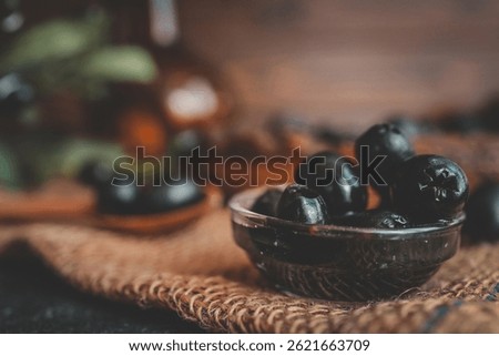 Rustic still life composition with black olives in a wooden bowl, a wooden spoon, olive branch, and glass bottle on burlap and dark table. Warm vintage food photography style.