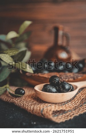 Rustic still life composition with black olives in a wooden bowl, a wooden spoon, olive branch, and glass bottle on burlap and dark table. Warm vintage food photography style.