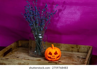 A rustic still life featuring a small, 3D-printed orange jack-o'-lantern and a glass jar of dried blue lavender on a vintage wooden tray against a textured magenta background.