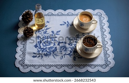 A rustic still life featuring two coffee cups, one filled with espresso and the other with coffee grounds, alongside a bottle of oil and pinecones on a blue floral doily.