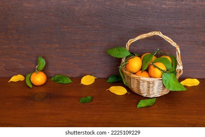 Rustic Still Life With Ripe Tangerines In A Wicker Basket And Green And Yellow Leaves On A Wooden Table. Harvest Time, Thanksgiving. Place For Text, Close-up, Front View, Copy Space, Mock Up