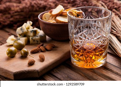 Rustic Still Life With Whiskey And Snacks. Crystal Glass With A Beautiful Pattern On A Wooden Table. Snack For Whiskey On Crumpled Paper Olives, Crackers, Cheese, Almonds, Pear, Apple.