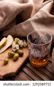 Rustic Still Life With Whiskey And Snacks. Crystal Glass With A Beautiful Pattern On A Wooden Table. Snack For Whiskey On Crumpled Paper Olives, Crackers, Cheese, Almonds, Pear, Apple.