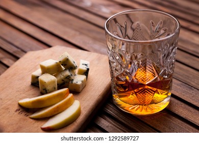 Rustic Still Life With Whiskey And Snacks. Crystal Glass With A Beautiful Pattern On A Wooden Table. Snack For Whiskey On Crumpled Paper Olives, Crackers, Cheese, Almonds, Pear, Apple.