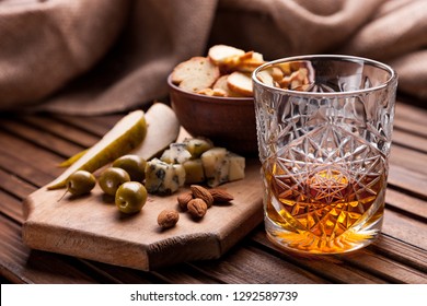 Rustic Still Life With Whiskey And Snacks. Crystal Glass With A Beautiful Pattern On A Wooden Table. Snack For Whiskey On Crumpled Paper Olives, Crackers, Cheese, Almonds, Pear, Apple.