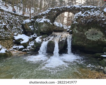 A rustic stone arch bridge spanning a small waterfall in a snowy forest. Water cascades over large rocks into a clear pool below, while patches of snow cover the bridge, ground, and surrounding boulders. The leafless trees and winter atmosphere create a picturesque blend of nature and old architecture, ideal for travel themes, seasonal concepts, and outdoor landscape designs.