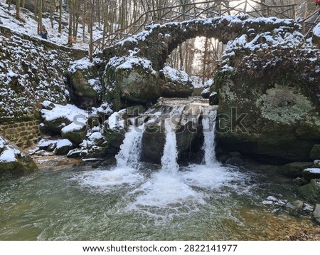 A rustic stone arch bridge spanning a small waterfall in a snowy forest. Water cascades over large rocks into a clear pool below, while patches of snow cover the bridge, ground, and surrounding boulders. The leafless trees and winter atmosphere create a picturesque blend of nature and old architecture, ideal for travel themes, seasonal concepts, and outdoor landscape designs.