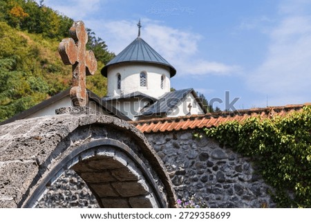 A rustic stone arch frames a quaint church and ivy-covered wall, set against a clear blue sky on a summer dunny day. Moraca Monastery, Kolasin, Montenegro