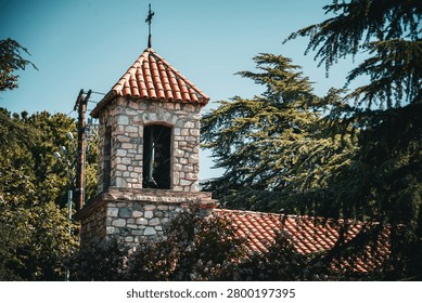 A rustic stone bell tower with a red tiled roof and a cross on top stands amidst lush green trees and foliage