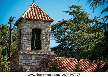 A rustic stone bell tower topped with a red tiled roof stands amidst lush green trees and purple flowers under a bright blue sky