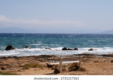 A rustic stone bench faces the vibrant blue Aegean Sea, set on a rocky shore under the clear summer sky. Waves crash gently against the rocks, while distant mountains fade into a hazy horizon. This tranquil seascape captures the serene, untamed beauty of coastal Greece, inviting moments of reflection and peaceful solitude.