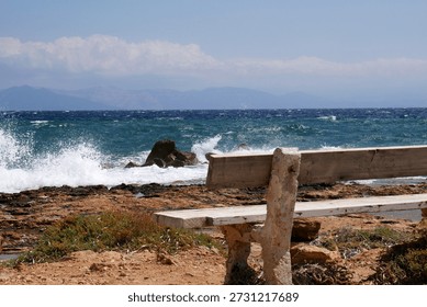 A rustic stone bench faces the vibrant blue Aegean Sea, set on a rocky shore under the clear summer sky. Waves crash gently against the rocks, while distant mountains fade into a hazy horizon. This tranquil seascape captures the serene, untamed beauty of coastal Greece, inviting moments of reflection and peaceful solitude.