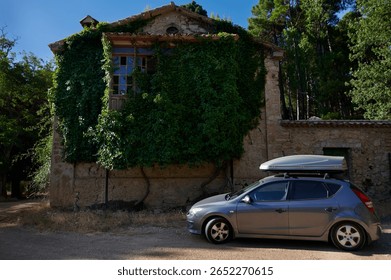 A rustic stone building partially covered in vibrant green ivy, set amid a tranquil forest. A compact silver car with a rooftop cargo box is parked in the foreground.