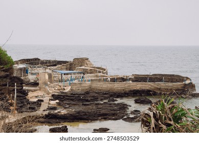 A rustic stone and concrete structure built into a dark, rocky volcanic coastline overlooking the sea. The scene features a small sheltered area with a blue roof, weathered steps, and natural coastal vegetation under a bright, overcast sky, reflecting a quiet and rugged maritime environment.