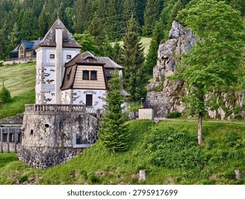 A rustic stone house with a small tower sits on a grassy riverside slope beneath dense evergreen forest. The scene shows weathered masonry walls, a plastered