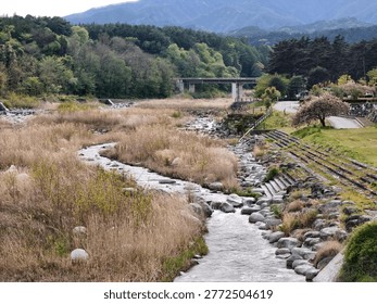 Rustic stone path winds through dry grassy fields. Riverbed stones line a tranquil, winding trail. Background features a bridge over calm waters. Mountains rise behind dense, lush green trees