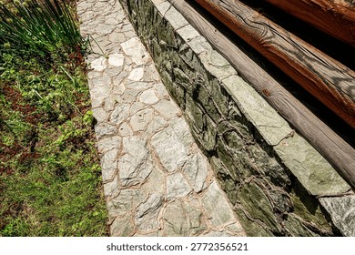 A rustic stone pathway bordered by a greenish-gray stone wall and weathered wooden beams. Lush greenery and wild plants flank the path, creating a natural, secluded ambiance.