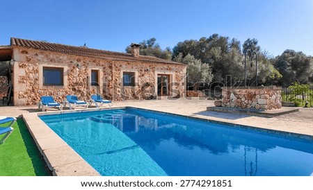 A rustic stone villa featuring a crystal-clear blue swimming pool, sun loungers, and a traditional water well under a bright blue sky