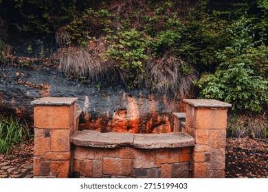 A rustic stone well structure built into a sandstone cliffside at the historic Ramlosa Brunnspark. The background shows wet rock stained orange by iron-rich mineral water, characteristic of this famou