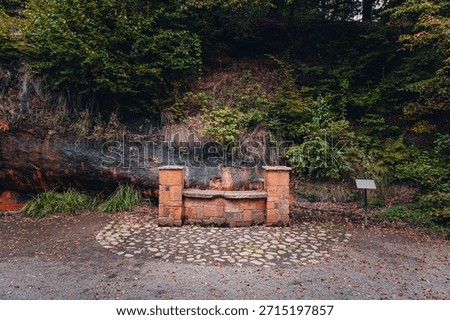 A rustic stone well structure built into a sandstone cliffside at the historic Ramlosa Brunnspark. The background shows wet rock stained orange by iron-rich mineral water, characteristic of this famou