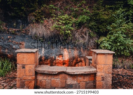 A rustic stone well structure built into a sandstone cliffside at the historic Ramlosa Brunnspark. The background shows wet rock stained orange by iron-rich mineral water, characteristic of this famou