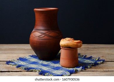 Rustic Style Pot And Mug From Clay With Milk And Sweet Brioche On Blue Napkin On Wooden Table, Black Background. Side View.