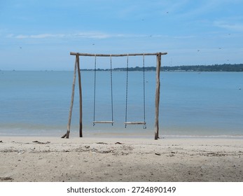 A rustic swing set on a sandy beach overlooking a calm sea under a clear blue sky.