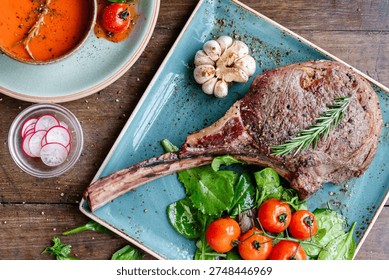 A rustic table setting with a grilled tomahawk steak, cherry tomatoes, garlic, and greens on a blue plate, accompanied by a bowl of soup and sliced radishes.