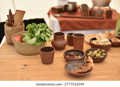 Rustic table setting with pottery dishes and fresh food.
Wooden table arranged with handmade clay cups, bowls and plates containing olives, nuts, bread and fresh vegetables. Traditional rustic dining scene with natural materials and artisan pottery.