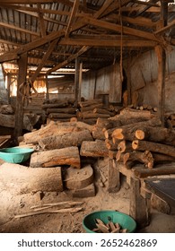 Rustic timber mart interior: rough logs piled under exposed wooden beams and corrugated roof. Natural light highlights textures, evoking raw materials, industry, and traditional woodworking.