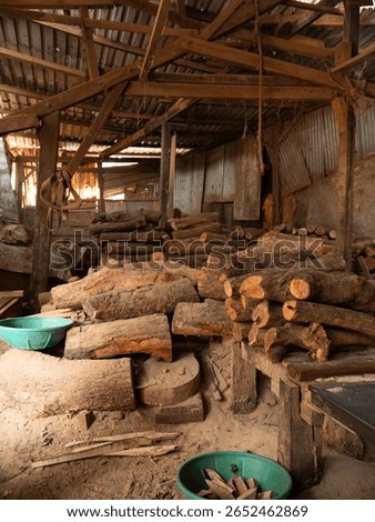 Rustic timber mart interior: rough logs piled under exposed wooden beams and corrugated roof. Natural light highlights textures, evoking raw materials, industry, and traditional woodworking.