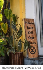 A rustic timber plaque requesting guests to unshoe leaning against a yellow wall next to polka dot begonia potted plants in a woven basket.
