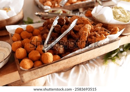 A rustic tray of crispy cheese balls, chicken tenders, and meatballs with tongs, set on a buffet table with a variety of complementary dishes in the background