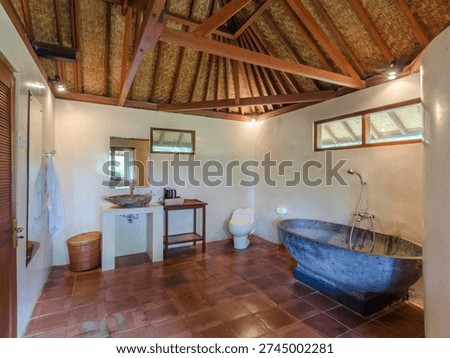 Rustic tropical bathroom interior featuring a natural stone bathtub, wooden ceiling beams, terracotta floor tiles, and minimalist sink area. Cozy cottage-style bathroom design with warm earthy tones and traditional craftsmanship details