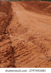 A rustic unpaved road featuring deep tire tracks carved into soft red clay earth under bright natural sunlight during daytime