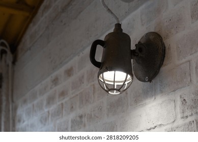 Rustic wall-mounted light fixture casts warm glow on brick wall. Vintage design features metal cage and handle. Perfect for cafes, lofts, or industrial-themed interiors