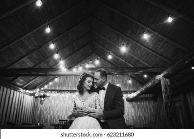 Rustic Wedding Couple Gently Hugging Under Retro Bulbs Lights In Wooden Barn. Space For Text. Newlyweds Embracing, Sensual Romantic Moment. Black White Photo