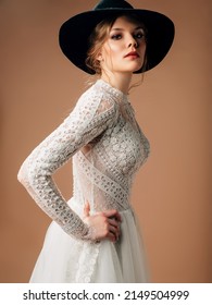 Rustic wedding dress. Elegant bridal gown with tender cotton lace, long transparent sleeves, lush tulle skirt. Beautiful young blonde lady bride with black cowboy hat supply. Studio shot.