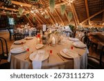 Rustic wedding reception setup with round tables, floral centerpieces, candles, string lights, hanging greenery, and elegant table settings in a wooden hall.