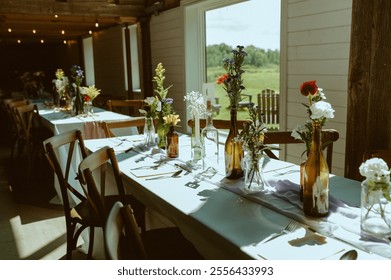 A rustic wedding reception table adorned with glass and amber vases filled with vibrant flowers, soft linens, and warm natural light streaming in through large windows.