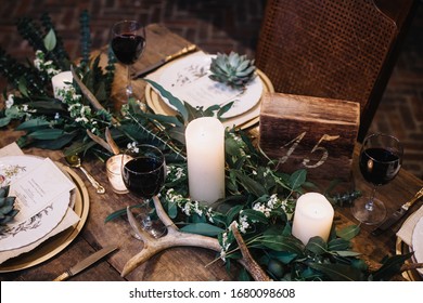 Rustic Wedding Table Seting With Vintage Plates, Green And White Eucalyptus Garland, Deer Antlers, Gold Utensils. Boho Style. Table Set For An Event, Party, Date Or Wedding. Red Wine Served