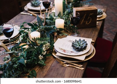Rustic Wedding Table Seting With Vintage Plates, Green And White Eucalyptus Garland, Deer Antlers, Gold Utensils. Boho Style. Table Set For An Event, Party, Date Or Wedding. Red Wine Served