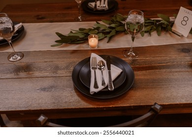 Rustic wedding table setting featuring a wooden table, black dinnerware, a greenery table runner, a soft candle centerpiece, and elegant glassware, creating a cozy and intimate ambiance.