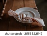 Rustic wedding table setting with a white plate, pink knotted napkin, and handwritten place card on a wooden table. Perfect for elegant, romantic, and modern event decor themes.