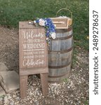 A rustic wedding welcome sign with blue and white flowers on a wooden barrel outdoors in Elkhart County, Indiana