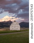 Rustic white barn in Bozeman, Montana, surrounded by tall grass, mountains and dramatic sunset day 