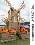 A rustic windmill model made of hay with pumpkins in wooden crates displayed in front of it. The windmill has a round body and four large blades. The pumpkins are of various sizes and colors.