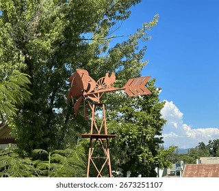 Rustic windmill weather vane with rooster silhouette against a clear blue sky and trees.