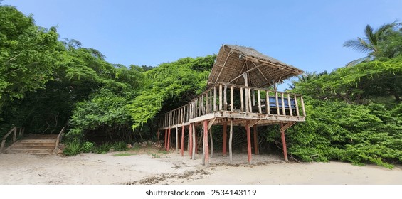 A rustic wooden beach hut elevated on stilts nestled against lush tropical greenery on a sandy beach. The open design offers a serene view of the tranquil coastline under a clear sky. - Powered by Shutterstock