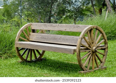 A rustic wooden bench with wagon wheels as supports, set on a lush green lawn with a backdrop of trees and foliage, creating a picturesque countryside setting - Powered by Shutterstock